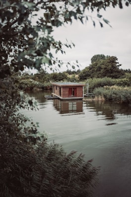 A small wooden houseboat floating on a calm river, surrounded by lush greenery and dense foliage. The overcast sky casts a muted light over the scene, enhancing the tranquil atmosphere.