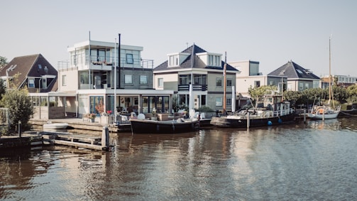 A serene waterfront scene with modern houses lined along the waterway. Boats are docked alongside the houses, and the area is surrounded by greenery. The architecture is contemporary, featuring large windows and clean lines, with a clear sky overhead.