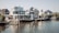 A serene waterfront scene with modern houses lined along the waterway. Boats are docked alongside the houses, and the area is surrounded by greenery. The architecture is contemporary, featuring large windows and clean lines, with a clear sky overhead.