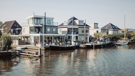 A serene waterfront scene with modern houses lined along the waterway. Boats are docked alongside the houses, and the area is surrounded by greenery. The architecture is contemporary, featuring large windows and clean lines, with a clear sky overhead.