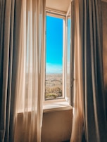 An open window with sheer curtains gently blowing, revealing a bright blue sky.