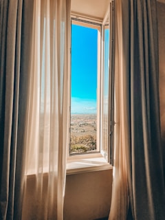 An open window with sheer curtains gently blowing, revealing a bright blue sky.