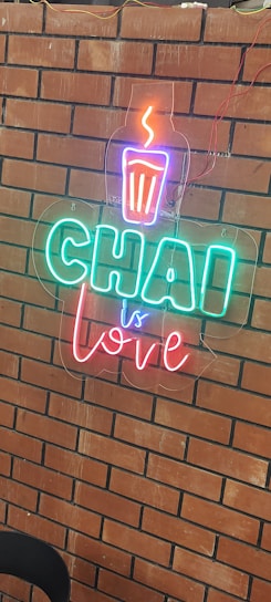 A brick wall adorned with a neon sign that features the phrase 'Chai is Love' in bright colors. The neon art includes a steaming cup symbol above the text.
