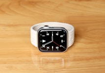 A modern smartwatch with a sleek black screen and a minimalist analog clock face featuring bold white hour and minute markings, set against a light-colored, woven strap. It rests on a smooth wooden surface.