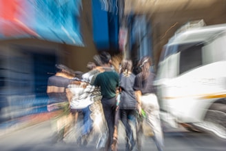 A blurred, dynamic scene featuring a group of people walking together on a street. The image captures movement, causing a streaked effect. To the right, a white vehicle is visible, and in the background, there are indistinct buildings with blue and red hues.