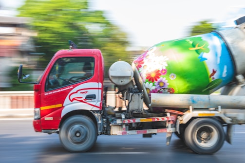 A red cement mixer truck is moving along a road. The truck has an eagle design on the side, and the drum is painted with colorful flowers and nature motifs. The background shows green foliage and blurred motion, indicating the vehicle is in motion.