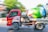 A red cement mixer truck is moving along a road. The truck has an eagle design on the side, and the drum is painted with colorful flowers and nature motifs. The background shows green foliage and blurred motion, indicating the vehicle is in motion.