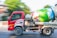 A red cement mixer truck is moving along a road. The truck has an eagle design on the side, and the drum is painted with colorful flowers and nature motifs. The background shows green foliage and blurred motion, indicating the vehicle is in motion.