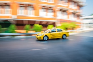 A yellow taxi is in motion on a street, with a blurred background of a building and greenery, indicating speed.