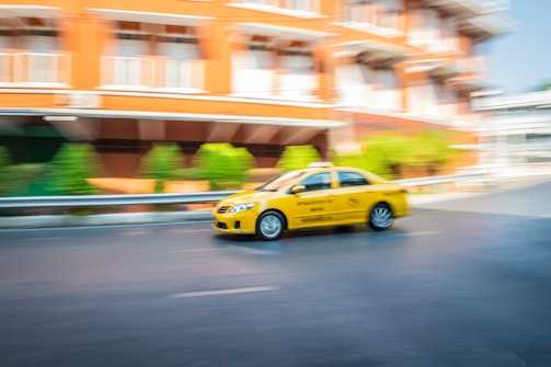 A yellow taxi is in motion on a street, with a blurred background of a building and greenery, indicating speed.