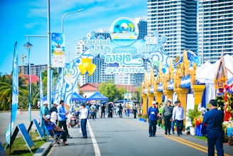 A vibrant outdoor festival scene featuring a decorative entrance gate with colorful banners and signs, indicating it's a sea festival. People are walking and gathering around, with some wearing badges and uniforms, likely those of event organizers. There are tents and booths set up along the street, adorned with blue and gold decorations. In the background, tall modern buildings rise against a clear blue sky.