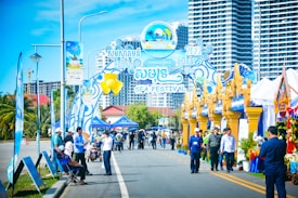 A vibrant outdoor festival scene featuring a decorative entrance gate with colorful banners and signs, indicating it's a sea festival. People are walking and gathering around, with some wearing badges and uniforms, likely those of event organizers. There are tents and booths set up along the street, adorned with blue and gold decorations. In the background, tall modern buildings rise against a clear blue sky.