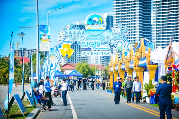 A vibrant outdoor festival scene featuring a decorative entrance gate with colorful banners and signs, indicating it's a sea festival. People are walking and gathering around, with some wearing badges and uniforms, likely those of event organizers. There are tents and booths set up along the street, adorned with blue and gold decorations. In the background, tall modern buildings rise against a clear blue sky.