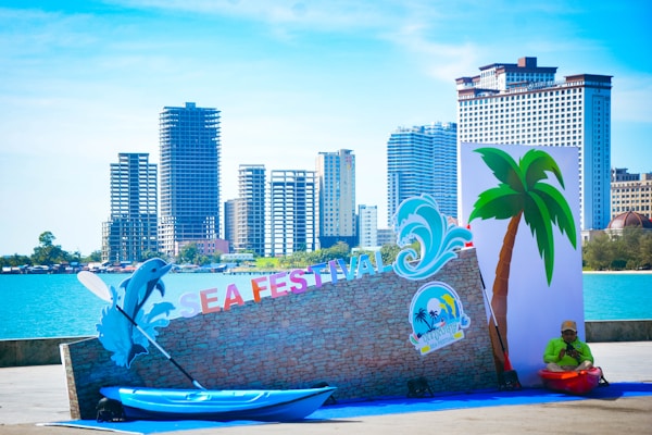 A coastal event setup includes a kayak placed by a decorative wall with the words 'SEA FESTIVAL' in colorful letters. A dolphin cutout and a wave graphic are part of the decoration. Tall buildings line the background, with clear blue water and a cloudless sky adding to the scene. A lone person in a bright outfit sits near the display.