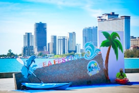 A coastal event setup includes a kayak placed by a decorative wall with the words 'SEA FESTIVAL' in colorful letters. A dolphin cutout and a wave graphic are part of the decoration. Tall buildings line the background, with clear blue water and a cloudless sky adding to the scene. A lone person in a bright outfit sits near the display.