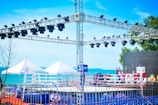A setup for an outdoor event with a boxing ring surrounded by flags and a metal truss structure equipped with multiple stage lights. The background features a body of water with clear blue skies, and there are white tents on the ground.