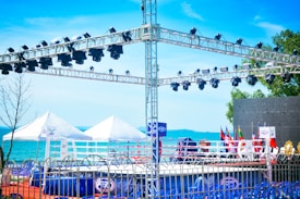A setup for an outdoor event with a boxing ring surrounded by flags and a metal truss structure equipped with multiple stage lights. The background features a body of water with clear blue skies, and there are white tents on the ground.