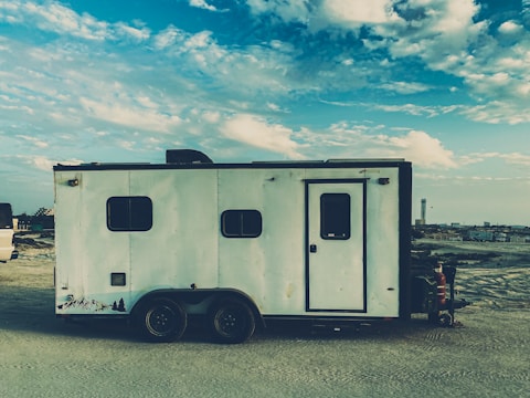 A large, rectangular white trailer with a minimalist design is parked in a barren, dusty area. The trailer features small windows and a door on its side. Above, a bright blue sky with scattered white clouds creates a serene backdrop.