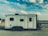 A large, rectangular white trailer with a minimalist design is parked in a barren, dusty area. The trailer features small windows and a door on its side. Above, a bright blue sky with scattered white clouds creates a serene backdrop.