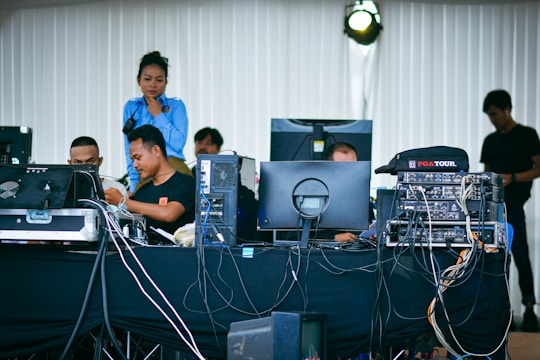 A group of people is working at a technical setup with multiple computer monitors and audio equipment. Cables and devices are scattered across the table, creating a scene of coordinated activity. The setting appears to be behind the scenes of an event, with individuals focused on their tasks, possibly in a control room or backstage area.