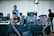 A group of people is working at a technical setup with multiple computer monitors and audio equipment. Cables and devices are scattered across the table, creating a scene of coordinated activity. The setting appears to be behind the scenes of an event, with individuals focused on their tasks, possibly in a control room or backstage area.