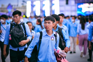 A group of people wearing blue uniforms, walking in an outdoor setting. Many are carrying backpacks, and some individuals are holding water bottles. The scene appears to be bustling with activity as people move together in the same direction.