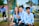 Three young people are sitting outdoors on a bench, wearing light blue shirts with a circular emblem. They appear to be smiling and enjoying a pleasant day in a park or campus setting. In the background, other people, also dressed in blue, are engaged in various activities.