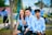 Three young people are sitting outdoors on a bench, wearing light blue shirts with a circular emblem. They appear to be smiling and enjoying a pleasant day in a park or campus setting. In the background, other people, also dressed in blue, are engaged in various activities.