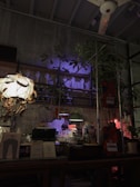 A dimly lit interior space features whimsical décor, including mannequin hands displayed on shelves and a mix of small plants. Ambient lighting casts colorful shadows and highlights, creating a mysterious and artistic atmosphere. The eclectic assembly of objects on the table includes lamps, jars, and various papers.