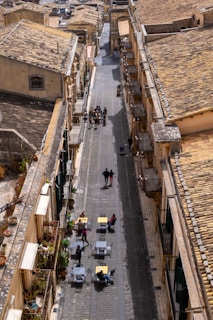 A narrow cobblestone street flanked by historic buildings featuring yellowish-brown tiled roofs. Outdoor seating with small tables and chairs is evident on one side, with a few people lounging and walking along the street. The scene is potentially urban and carries a cozy, casual vibe, enhanced by potted plants and balconies.