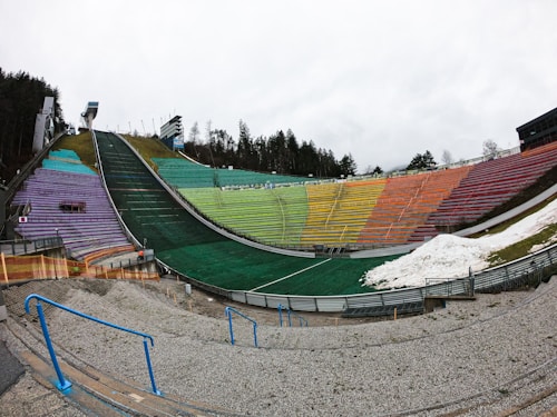 A ski jumping hill with colored seating areas divided into multiple colors including green, yellow, orange, and purple. There is a large ramp in the center. Snow is visible on one side of the seating area. The background includes a forested area with trees and a cloudy sky.