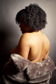 A person with curly hair is seen from the back, partially wrapped in a luxurious fur garment. The lighting accentuates the texture of the hair and the softness of the fur, creating a contrast with the smooth skin.