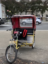 A bright yellow pedicab with a red canopy is parked on a city street, with a sign reading 'Central Park' attached to the front. The background includes a busy street scene with vehicles, trees, and a fountain visible.