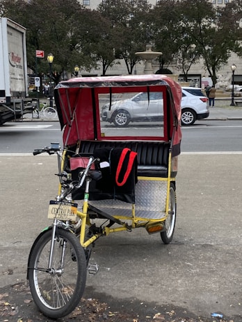 A bright yellow pedicab with a red canopy is parked on a city street, with a sign reading 'Central Park' attached to the front. The background includes a busy street scene with vehicles, trees, and a fountain visible.
