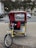 A bright yellow pedicab with a red canopy is parked on a city street, with a sign reading 'Central Park' attached to the front. The background includes a busy street scene with vehicles, trees, and a fountain visible.