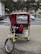 A bright yellow pedicab with a red canopy is parked on a city street, with a sign reading 'Central Park' attached to the front. The background includes a busy street scene with vehicles, trees, and a fountain visible.