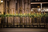 Ornate black metal railings are decorated with a garland of greenery and small lights. Behind the railings, there are brass luggage carts arranged in a row. The setting is likely a hotel entrance, featuring a stone facade with large windows.