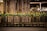 Ornate black metal railings are decorated with a garland of greenery and small lights. Behind the railings, there are brass luggage carts arranged in a row. The setting is likely a hotel entrance, featuring a stone facade with large windows.