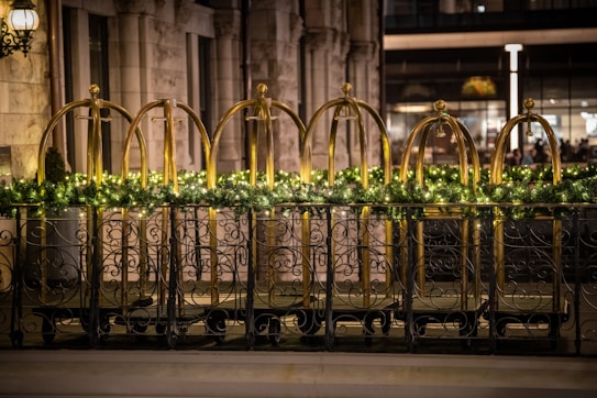 Ornate black metal railings are decorated with a garland of greenery and small lights. Behind the railings, there are brass luggage carts arranged in a row. The setting is likely a hotel entrance, featuring a stone facade with large windows.