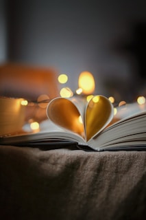 An open book with its pages curved inward to form the shape of a heart, set against a background illuminated by warm, soft fairy lights that create a bokeh effect.