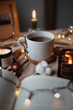 A cozy scene featuring a white mug of coffee placed on a wooden coaster surrounded by soft, warm lighting from candles and string lights. Cotton bolls and a small red winter-themed figure are arranged nearby, adding a touch of homeliness. An open book is partially visible in the foreground, enhancing the relaxed and inviting atmosphere.