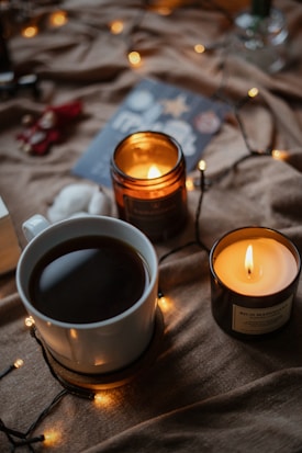A cozy setting with warm, ambient lighting featuring a white mug filled with a dark beverage, possibly coffee or tea, placed on a wooden coaster. Nearby, two brown candles are lit, emitting a soft, glowing light. The background includes a blurred-out card with festive decorations, and string lights are scattered across a textured, brown fabric.