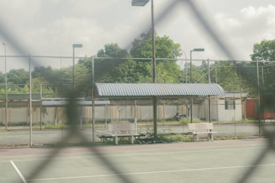 A fenced tennis court is visible with a wire fence in the foreground. Beyond the court, there's a corrugated metal roof structure with some benches and greenery. The sky is slightly overcast with visible clouds, lending a calm ambiance.