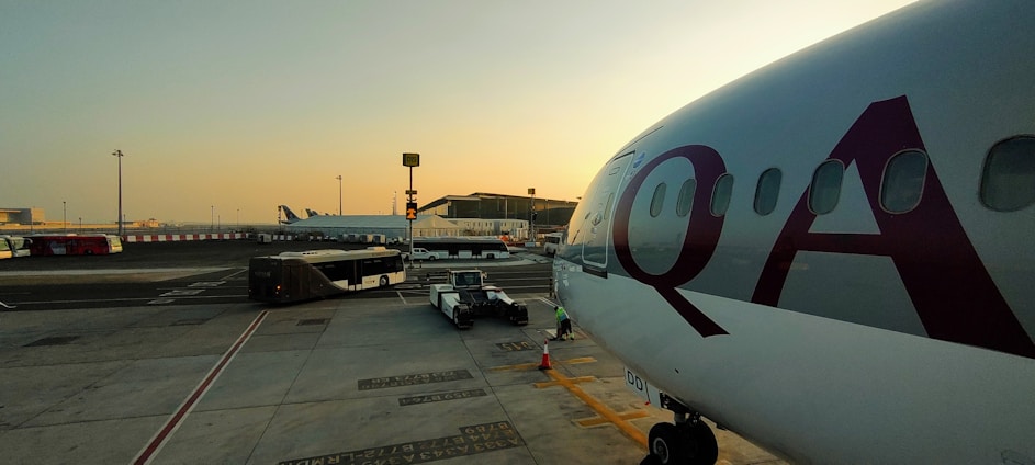 A large passenger airplane is parked at an airport gate with the letters 'QA' visible on the fuselage. The tarmac is busy with ground support vehicles, including a bus and luggage carts, moving around. In the background, there is a terminal building with various signs and infrastructure. The sky is clear with a warm, early morning or late afternoon light.