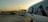 A large passenger airplane is parked at an airport gate with the letters 'QA' visible on the fuselage. The tarmac is busy with ground support vehicles, including a bus and luggage carts, moving around. In the background, there is a terminal building with various signs and infrastructure. The sky is clear with a warm, early morning or late afternoon light.