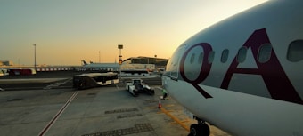 A large passenger airplane is parked at an airport gate with the letters 'QA' visible on the fuselage. The tarmac is busy with ground support vehicles, including a bus and luggage carts, moving around. In the background, there is a terminal building with various signs and infrastructure. The sky is clear with a warm, early morning or late afternoon light.