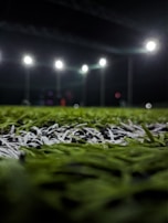 A close-up perspective of artificial grass with bright stadium lights illuminating from above. The focus is primarily on the green and white blades of the synthetic turf, with the background showing blurred outlines of light fixtures and possible figures.