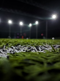 Close-up of lush green artificial turf with cricket ball and bat resting on it under bright floodlights.