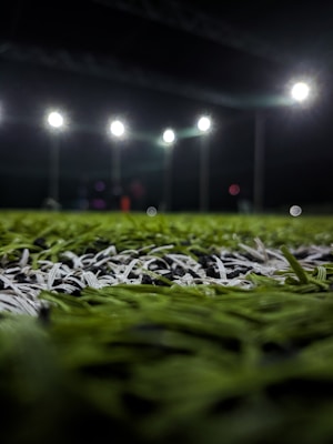 A close-up perspective of artificial grass with bright stadium lights illuminating from above. The focus is primarily on the green and white blades of the synthetic turf, with the background showing blurred outlines of light fixtures and possible figures.