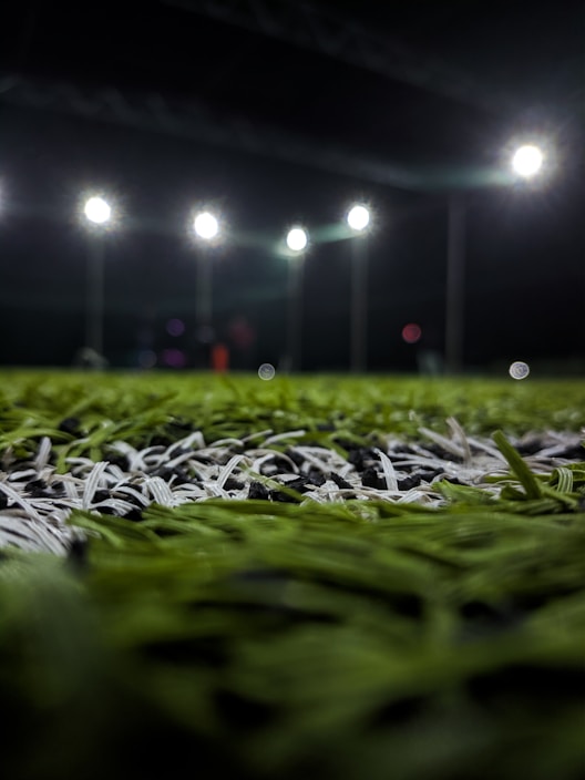 A close-up perspective of artificial grass with bright stadium lights illuminating from above. The focus is primarily on the green and white blades of the synthetic turf, with the background showing blurred outlines of light fixtures and possible figures.